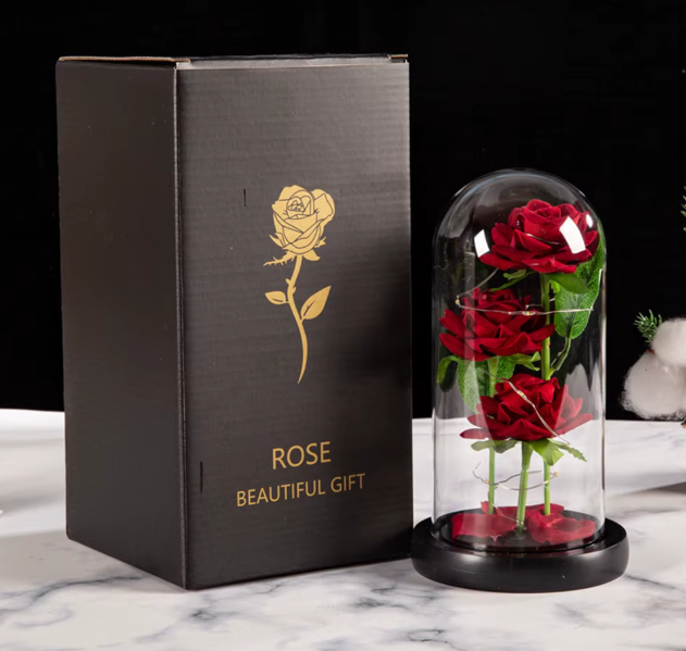 Black gift box with gold rose design and red roses in a glass dome on a marble surface.